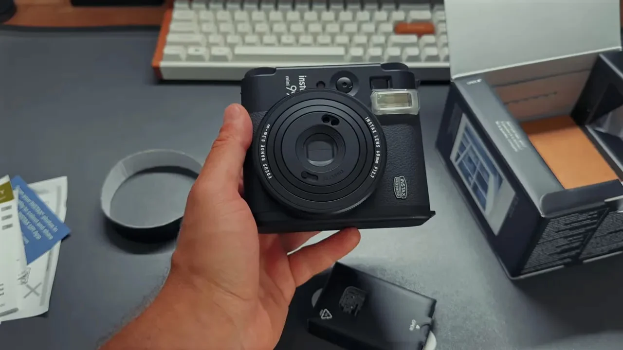 Front view of Instax Mini 99 held in one hand showing lens, flash, and front controls