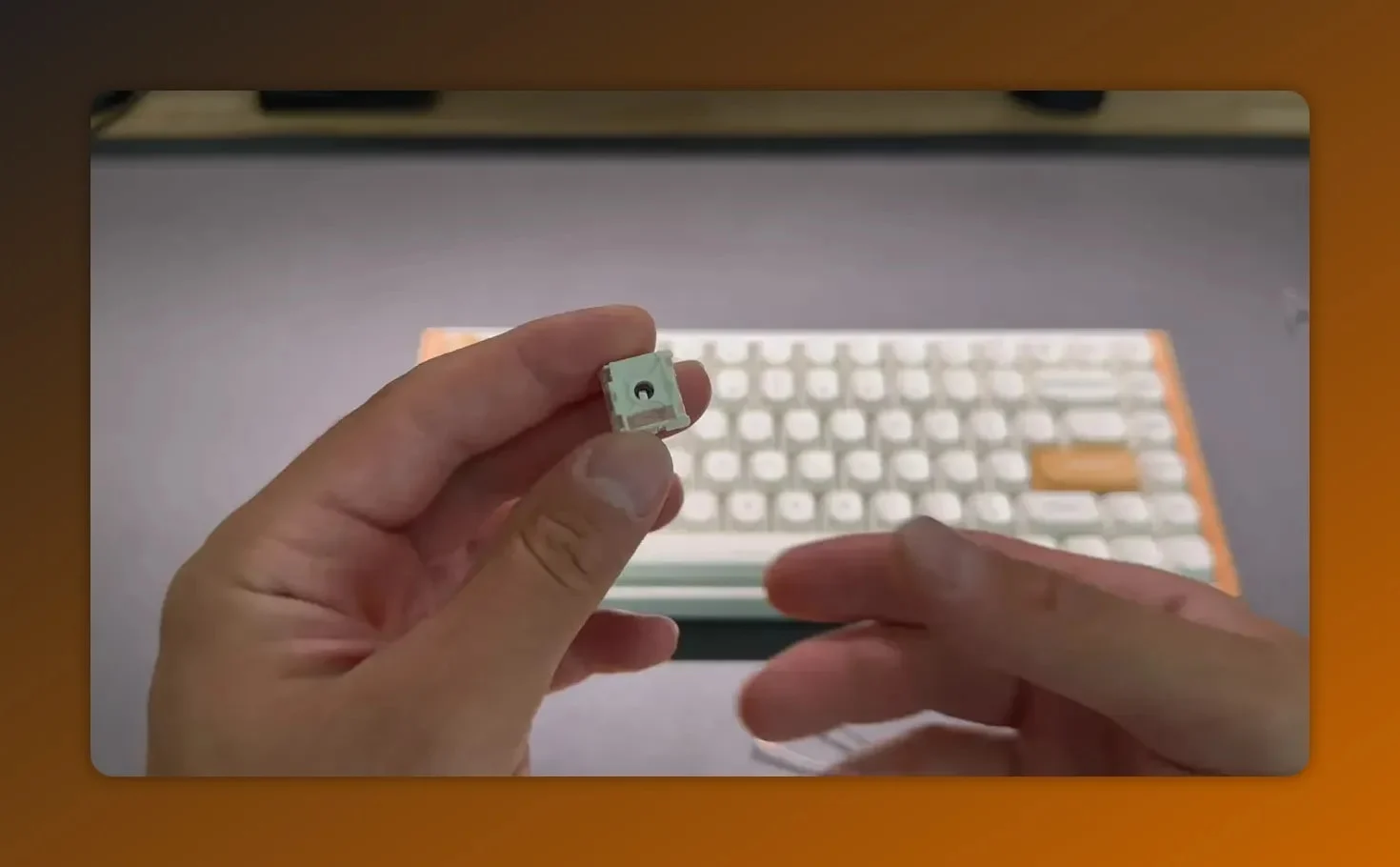 Close-up of a Keychron HE magnetic switch held between fingers showing the bottom and stem, with the keyboard blurred in the background.